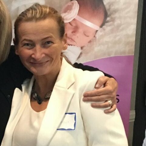Beata, a Tampa Postpartum Doula smiles at the camera in a white blazer. Behind her is a large poster featuring a sleeping newborn with a white headband. The atmosphere is warm and professional, likely at a health or maternity expo in Tampa Bay.