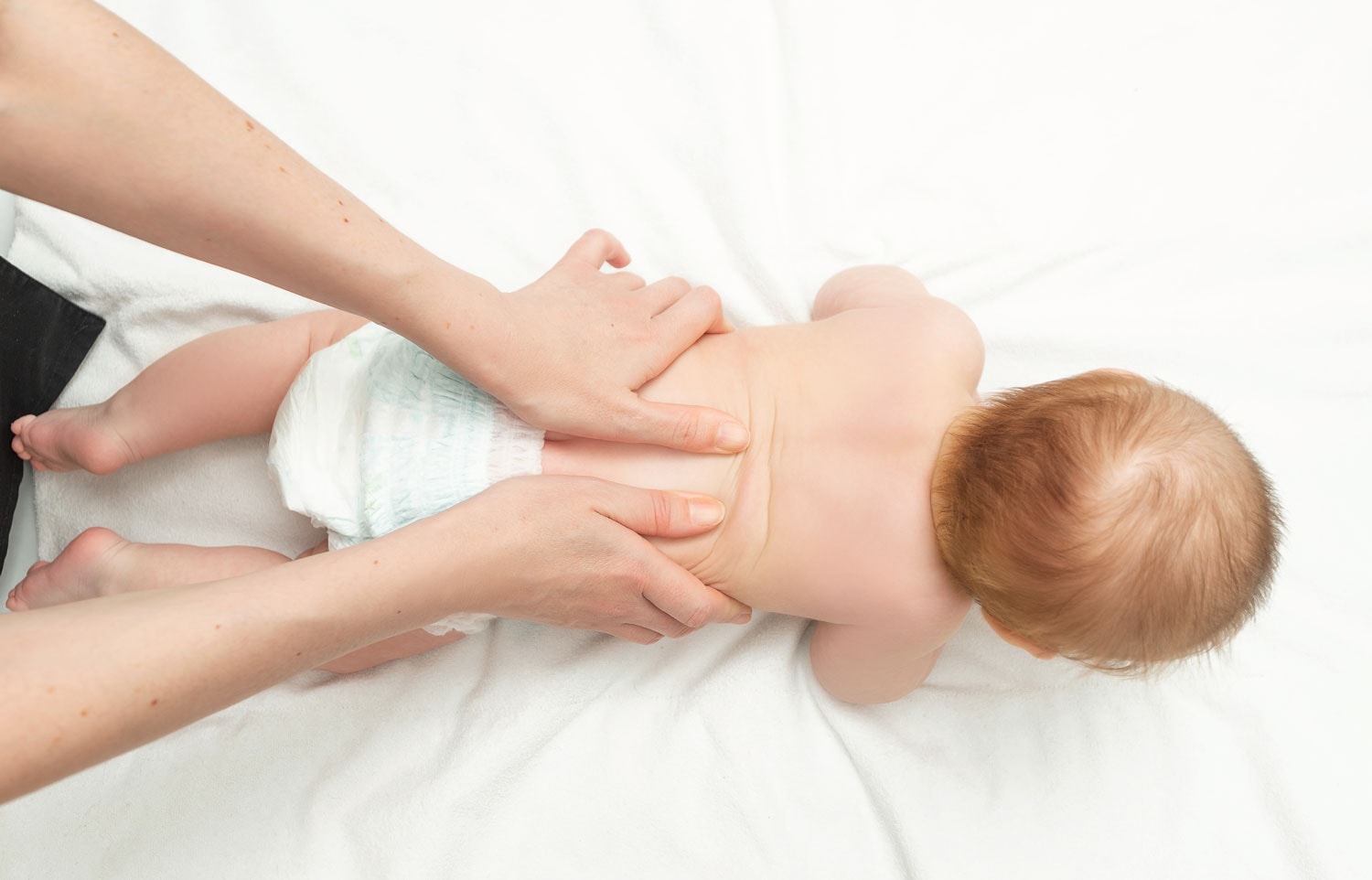 An infant receiving a gentle back massage from a Postpartum Doula's hands, lying comfortably on a white blanket, highlighting the importance of touch in baby's growth and relaxation.
