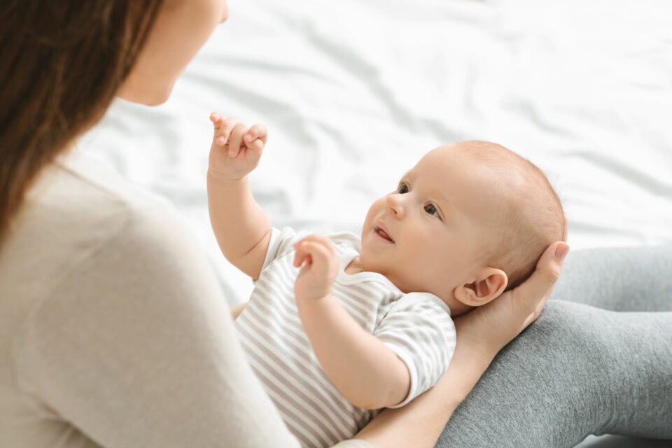 An infant with bright, attentive eyes lies cradled in the lap of a caregiver, looking up with a look of trust and wonder, set against a soft, white bedspread background.