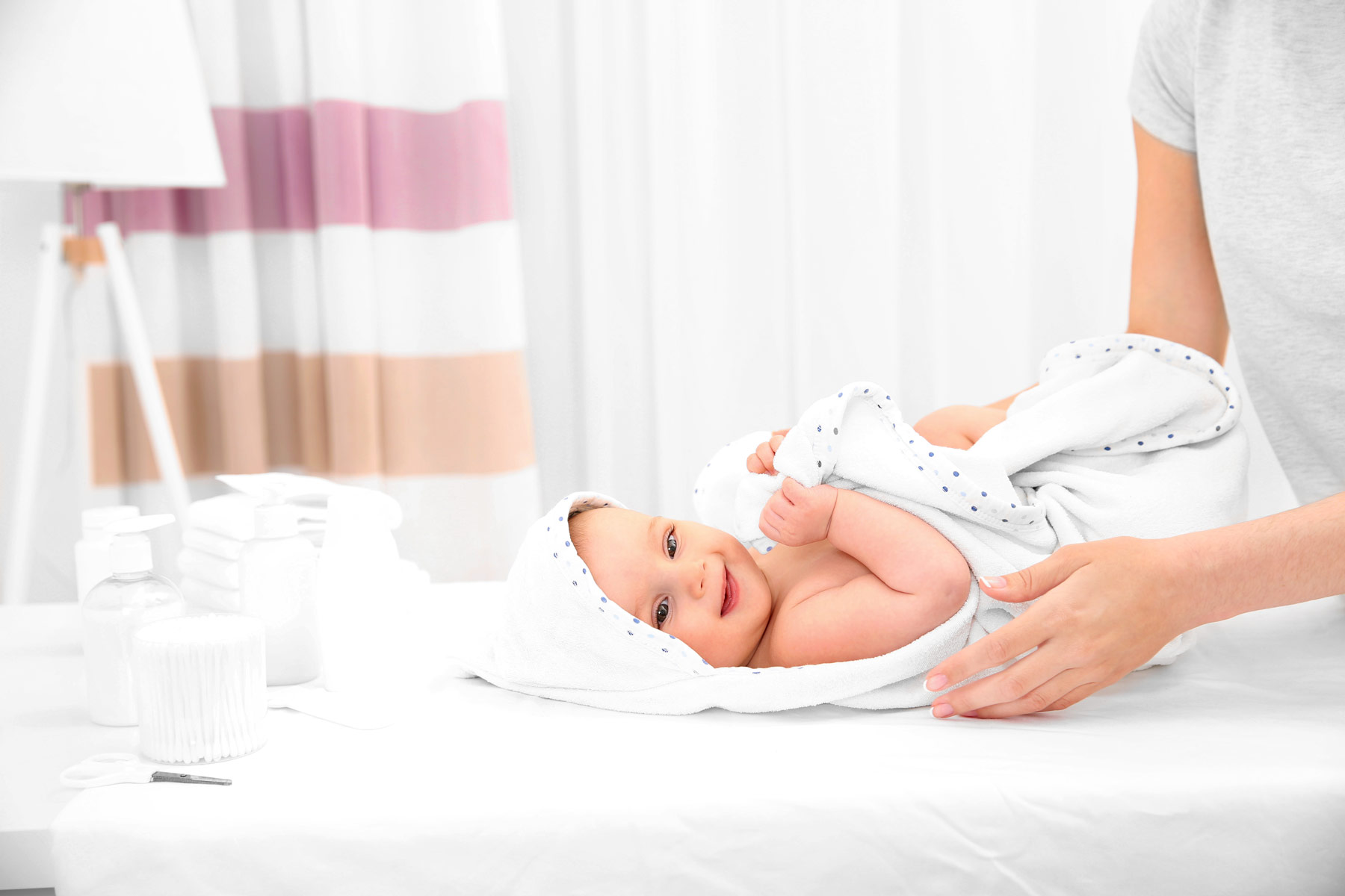 A content baby wrapped in a soft, white blanket with polka dots, smiling and gazing upwards, being gently cradled by a caregiver's hands on a changing table, surrounded by baby care products.
